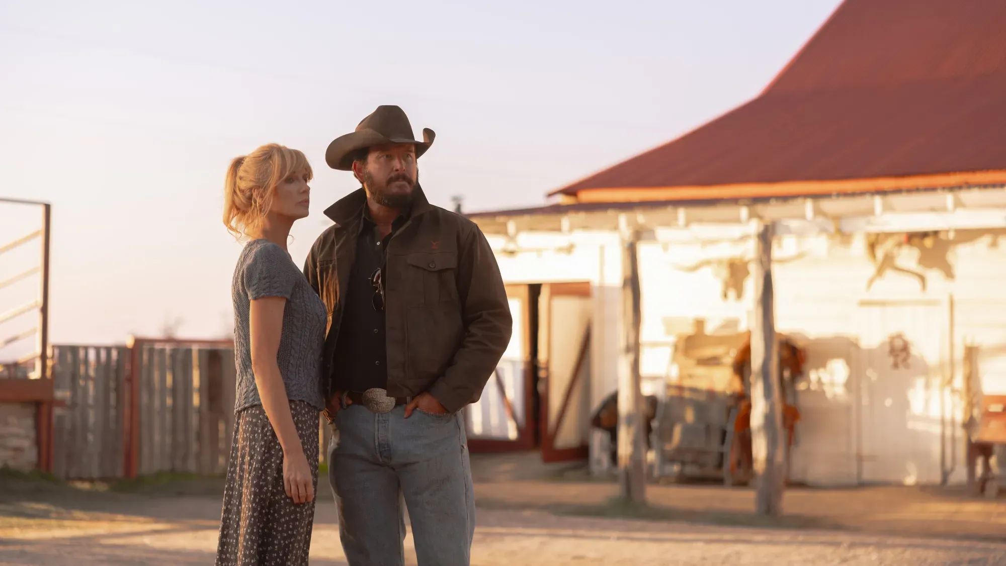 Beth and Rip Wheeler stand together outside the ranch buildings at golden hour, surveying their Texas territory