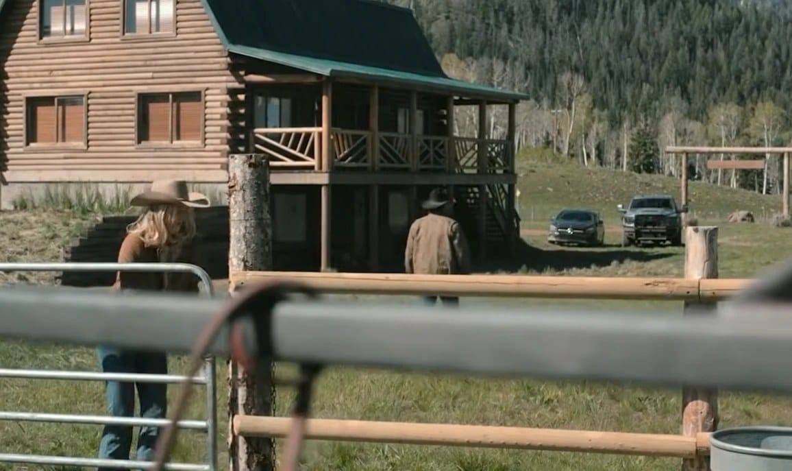 A log ranch house with a person in a cowboy hat at a gate and trucks parked beyond it.