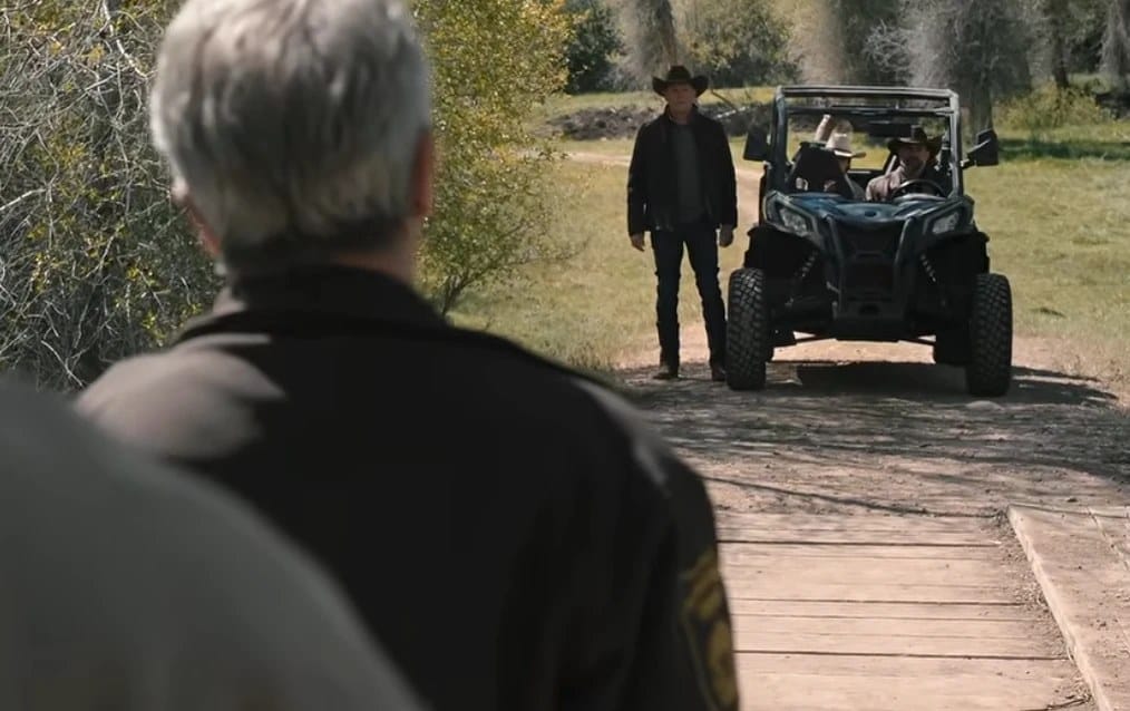 A man in a sheriff's jacket faces a man standing in front of an approaching utility vehicle.
